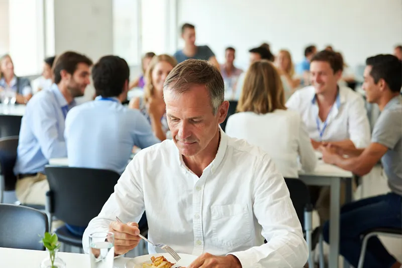 Un homme en train de déjeuner seul à la cantine pendant que ses collègues déjeunent ensemble 